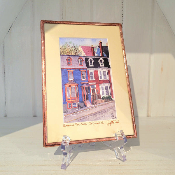 Copper framed artwork titled "Garrison Heritages" by Cynthia Noel of colorful houses on a stand against a white background