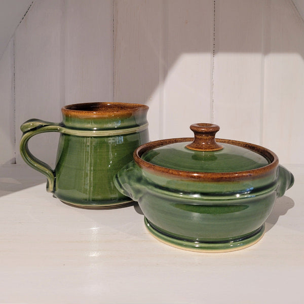 Two green ceramic items, a cream/milk jug and a sugar pot with lid, on a white surface with a wooden panel background.