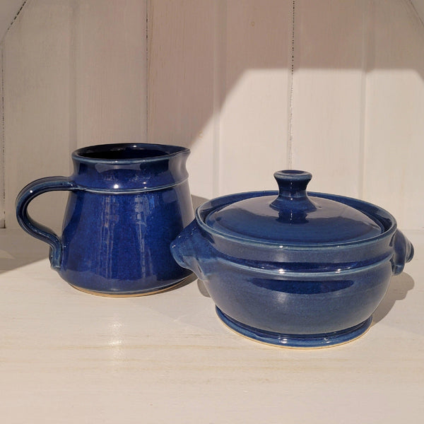 Two blue ceramic items, a cream/milk jug and a sugar pot with lid, on a white surface with a wooden panel background.