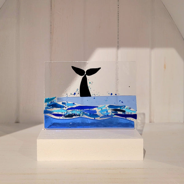 Decorative fused glass piece with a whale tail placed on a white base against a neutral background