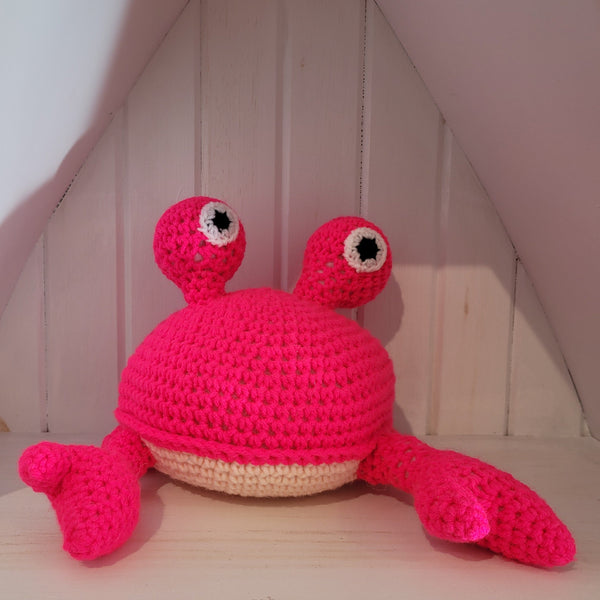 Crocheted pink crab stuffed toy on a wooden surface with a white wall background
