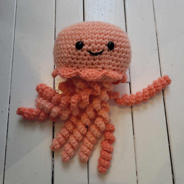 peach-coloured crochet plush toy jellyfish with smiley face and colourful tentacles on a white background